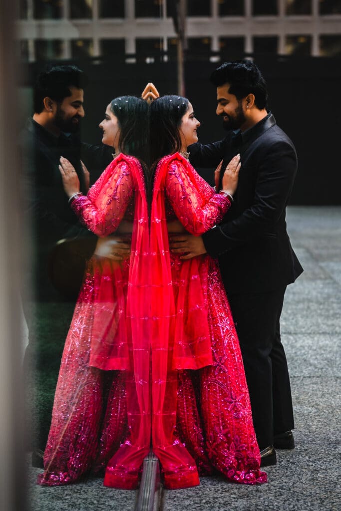South Asian couple posing in downtown Toronto beside a reflective glass wall, creating a mirrored double image—bride in a bright red embellished lehenga with dupatta and groom in a black suit, standing close and smiling in an intimate, cinematic portrait.
