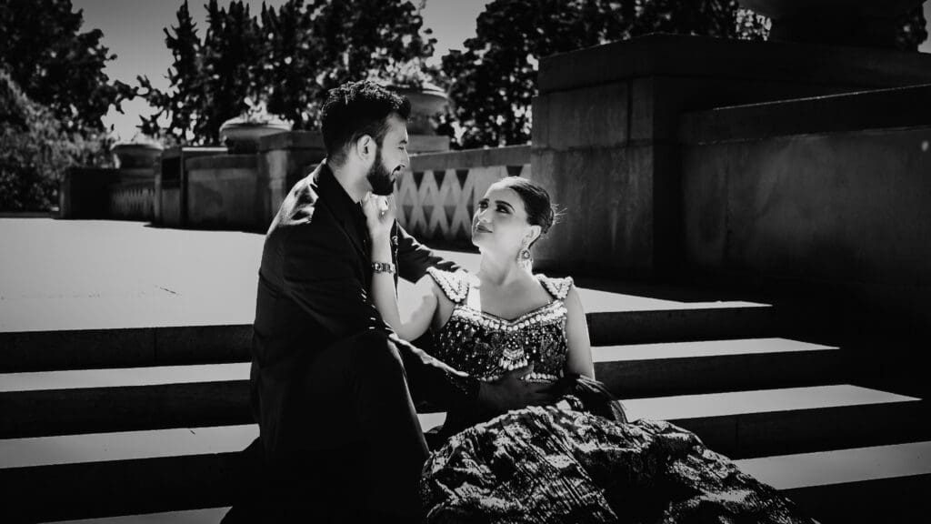Black and white Parkwood Estate wedding portrait of a South Asian couple sitting on stone steps, bride in embellished lehenga looking up at groom.