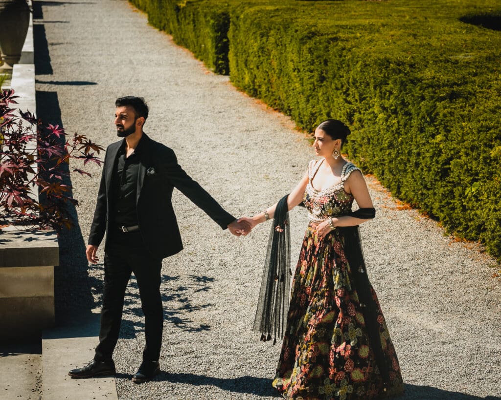 outh Asian wedding couple walking hand in hand at Parkwood Estate, Oshawa — editorial garden portrait with manicured hedges