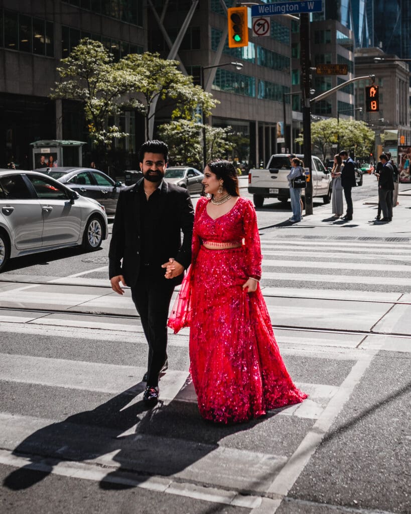 South Asian couple holding hands and walking across a downtown Toronto crosswalk on Wellington St W, bride in a red sequined lehenga and groom in a black suit, captured in bright natural light with city buildings and traffic in the background.