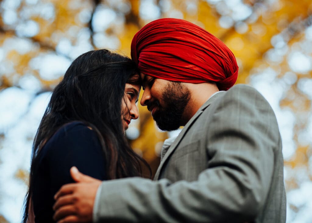 Close-up engagement portrait of a Sikh couple embracing, groom in red turban and grey suit, captured outdoors in Montreal with autumn bokeh.
