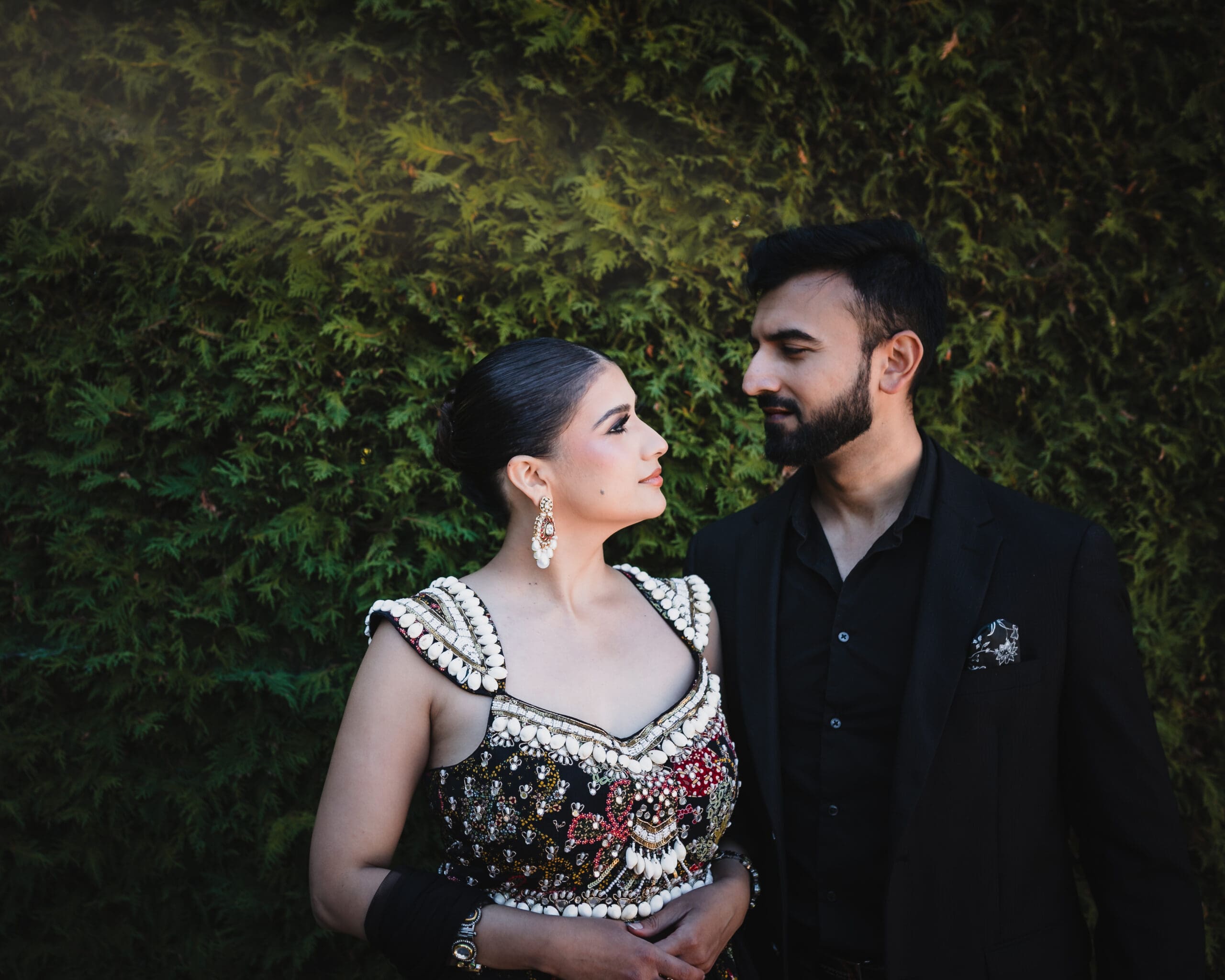 Wedding couple portrait in front of evergreen greenery at Parkwood Estate in Oshawa, Ontario, bride in embellished lehenga and groom in black suit sharing an intimate moment, cinematic South Asian wedding photography near Toronto and GTA.