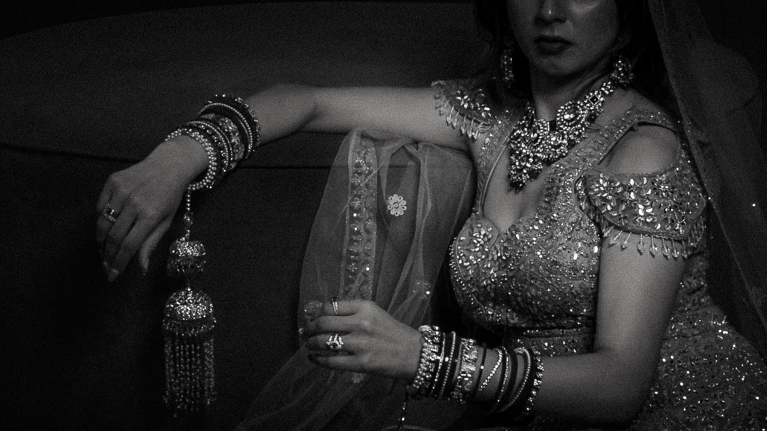 Black and white South Asian bridal portrait of a bride in an embellished lehenga, wearing statement necklace and bangles with kalire, seated in a moody editorial pose.