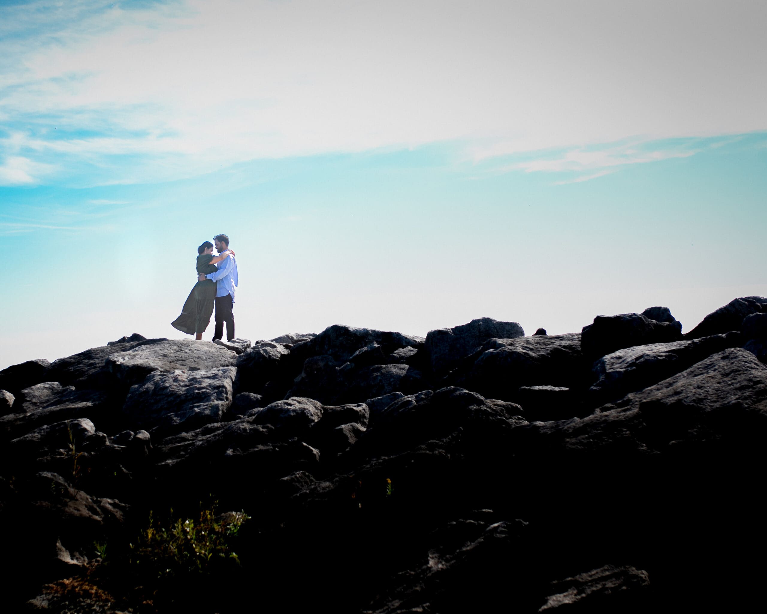 Couple embracing on the rocky waterfront at Colonel Samuel Smith Park in Toronto during a pre-wedding photo session, framed against an open blue sky.