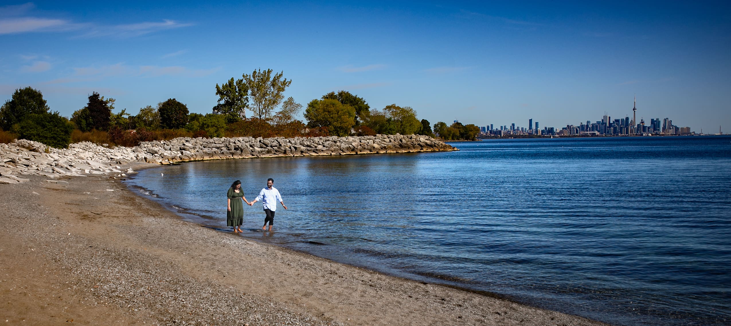 Pre-wedding couple walking barefoot along the shoreline at Colonel Samuel Smith Park in Toronto, with Lake Ontario and the Toronto skyline in the distance.