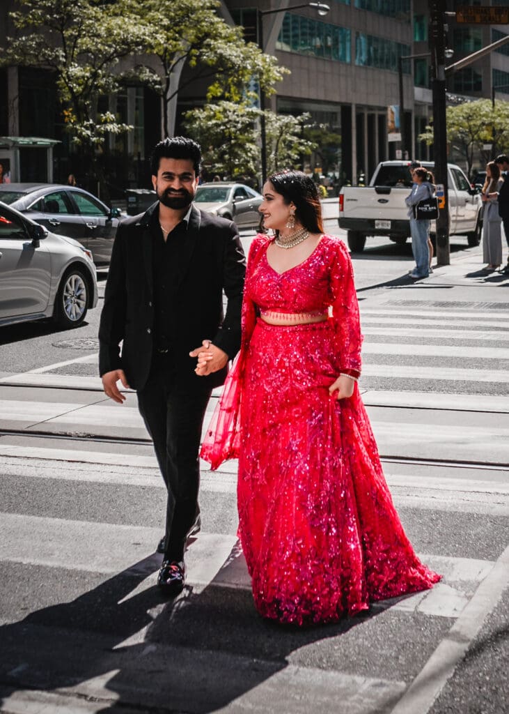 South Asian wedding couple walking downtown Toronto, bride in a red lehenga looking at groom in a black suit, candid street portrait with city light and movement
