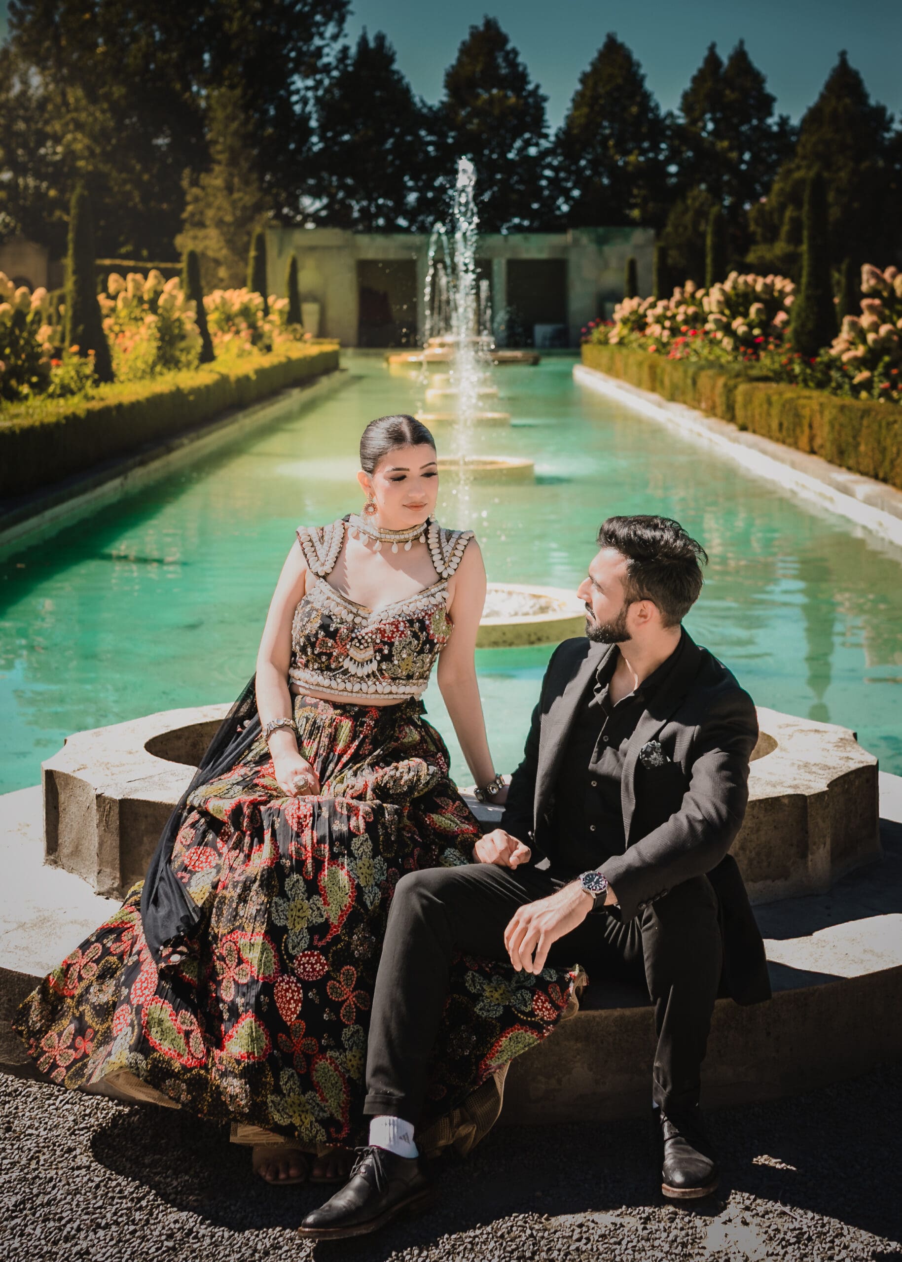 Wedding couple sitting by the long fountain pool at Parkwood Estate in Oshawa, Ontario, bride in embroidered lehenga and groom in black suit, cinematic estate garden wedding photography near Toronto and GTA.