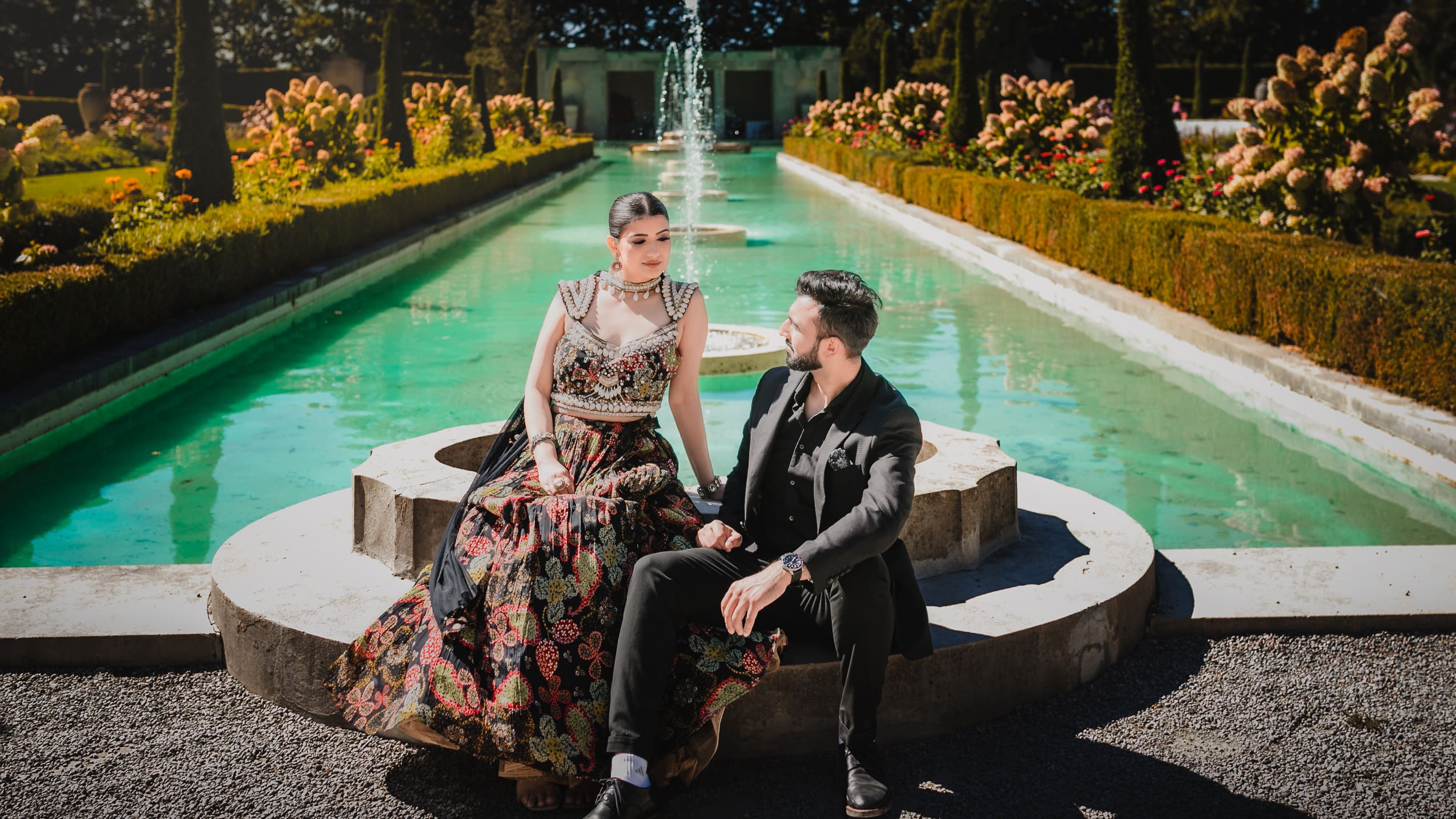 South Asian pre-wedding couple sitting by the long fountain pool at Parkwood Estate in Oshawa, Ontario, bride in embroidered lehenga and groom in black suit, cinematic garden portrait near Toronto and GTA.