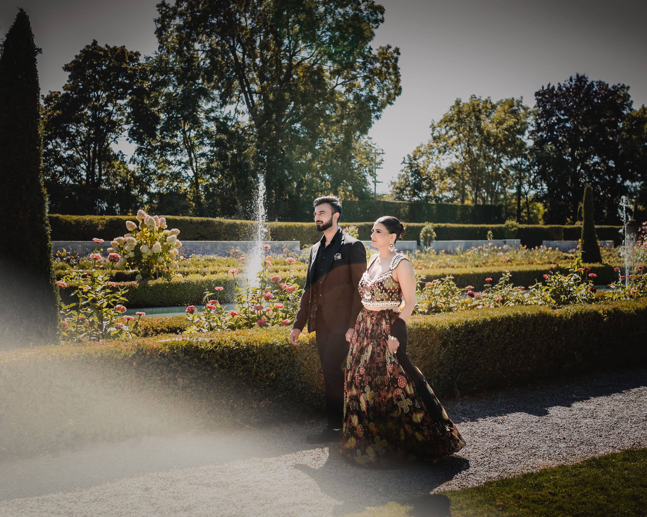 Wedding couple walking through the formal gardens at Parkwood Estate in Oshawa, Ontario, bride in embroidered lehenga and groom in black suit with fountain and flowers behind them, cinematic South Asian wedding photography near Toronto and GTA.