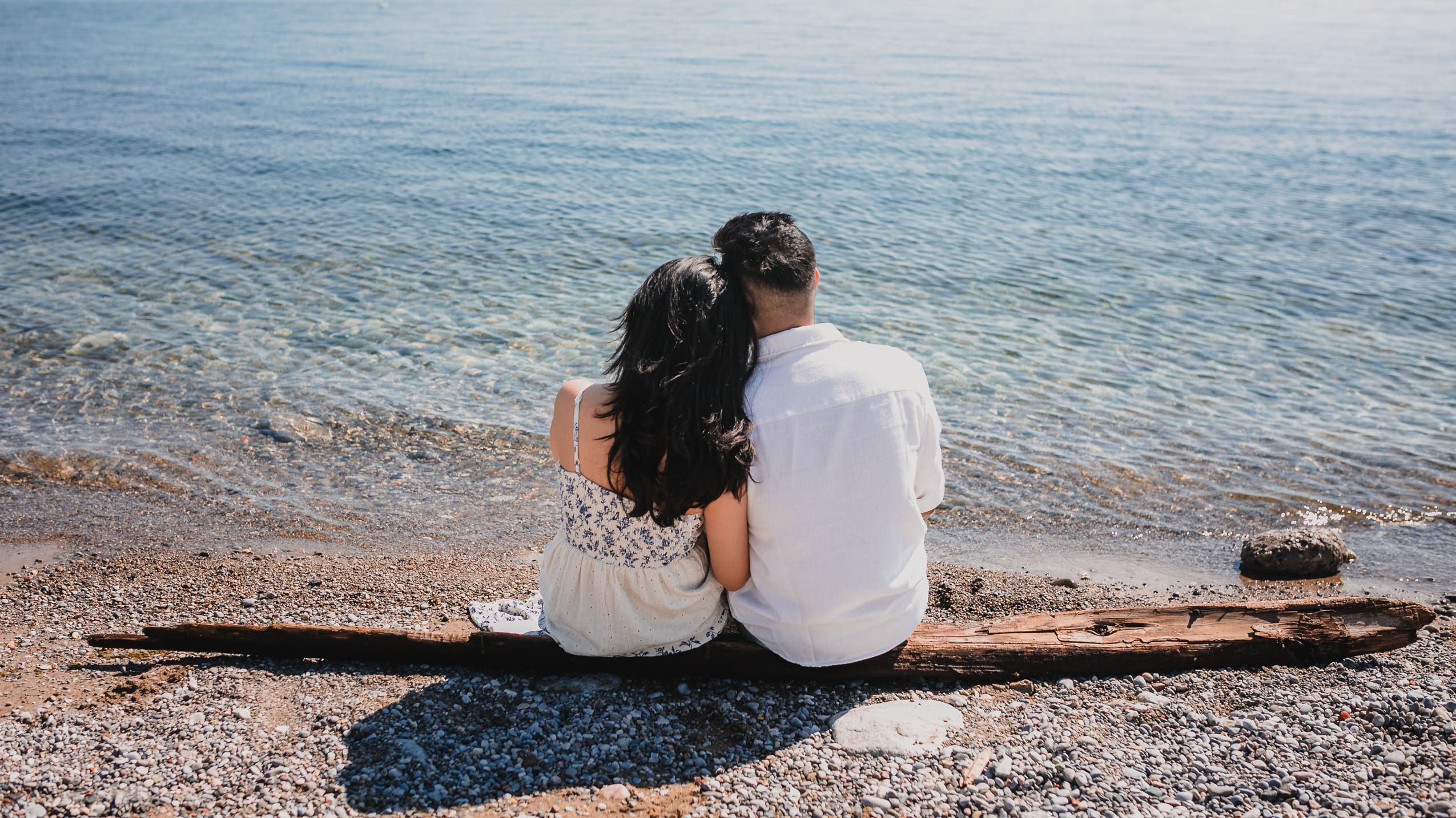 Couple sitting close together on a driftwood log at Scarborough Bluffs, looking out over calm Lake Ontario during a relaxed candid photo session.