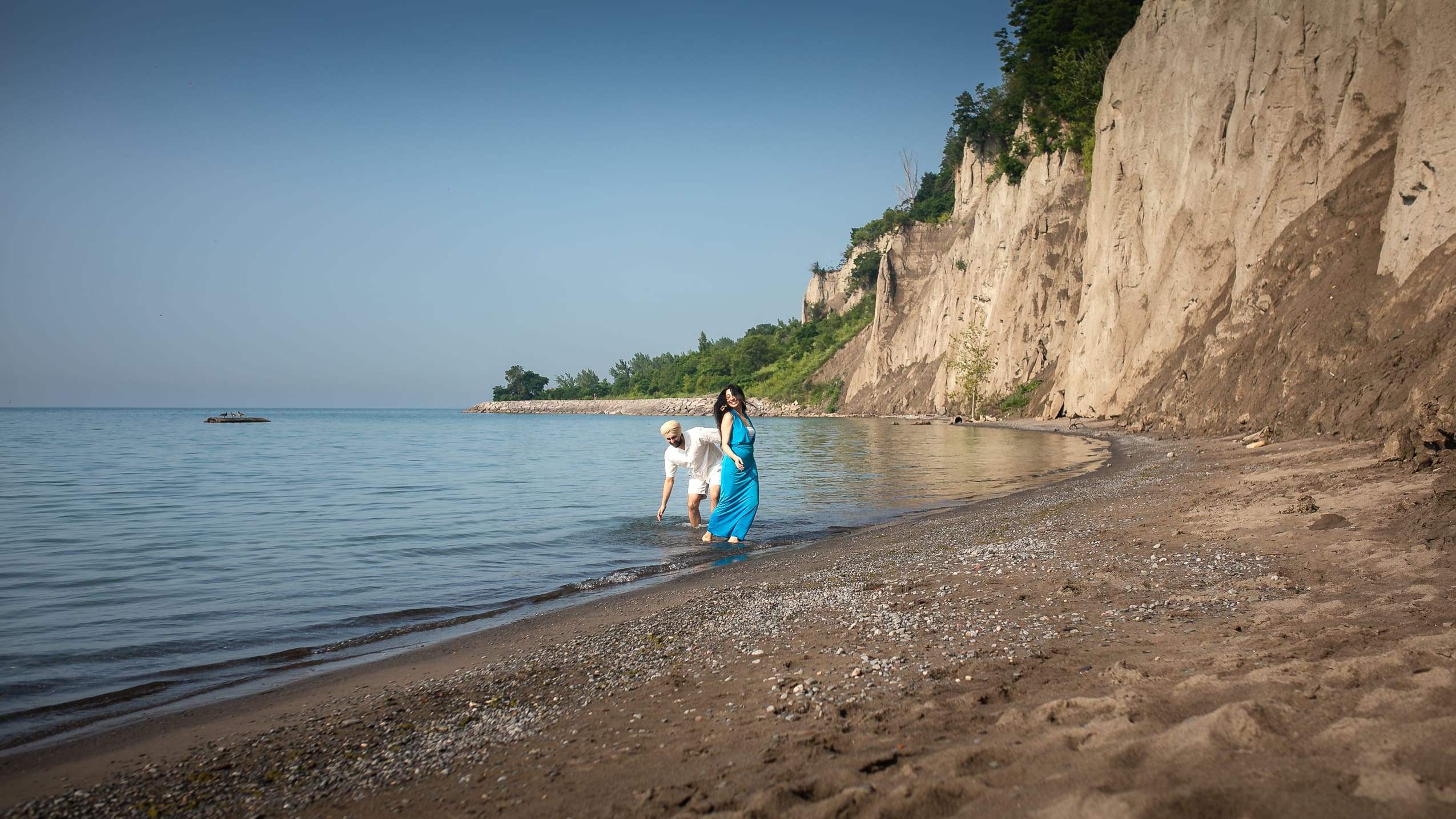 Couple walking barefoot along the shoreline at Scarborough Bluffs during a pre-wedding photoshoot in Toronto, with Lake Ontario and the cliffs in the background.