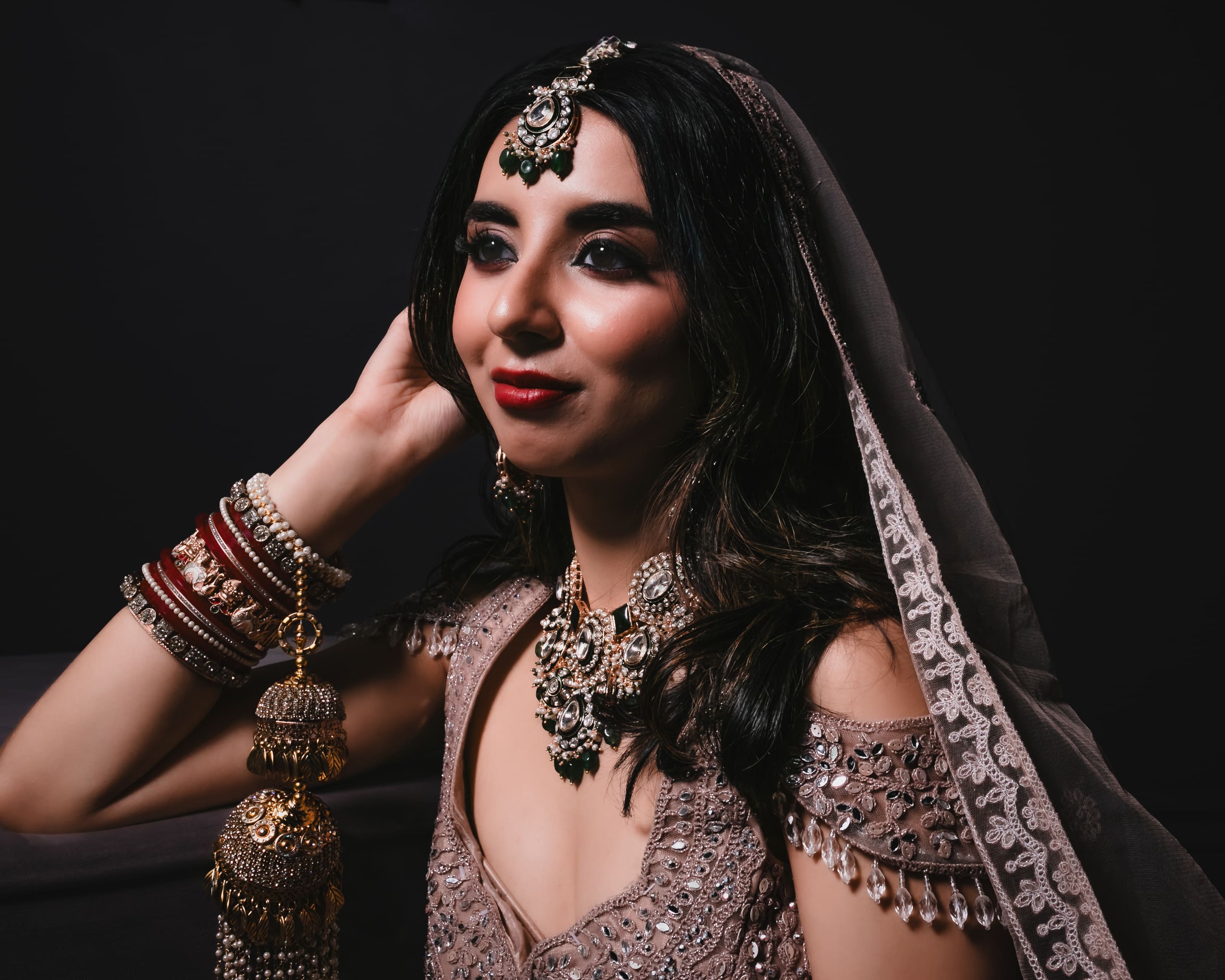 South Asian bridal editorial portrait in embellished outfit and veil, wearing kundan necklace, earrings, and maang tikka under dramatic studio lighting.