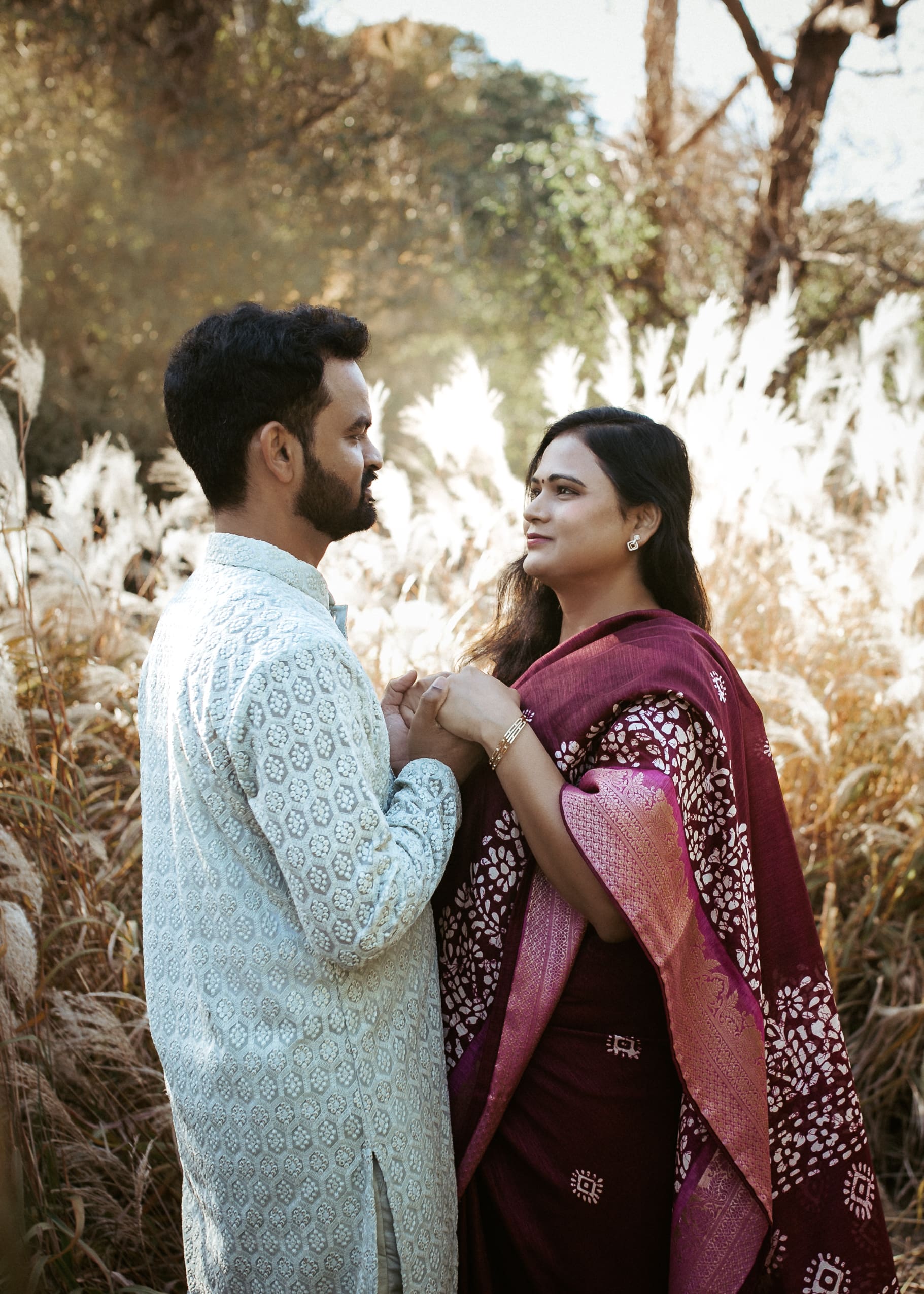 South Asian couple portrait in traditional attire, holding hands in tall golden grass during a warm outdoor session.
