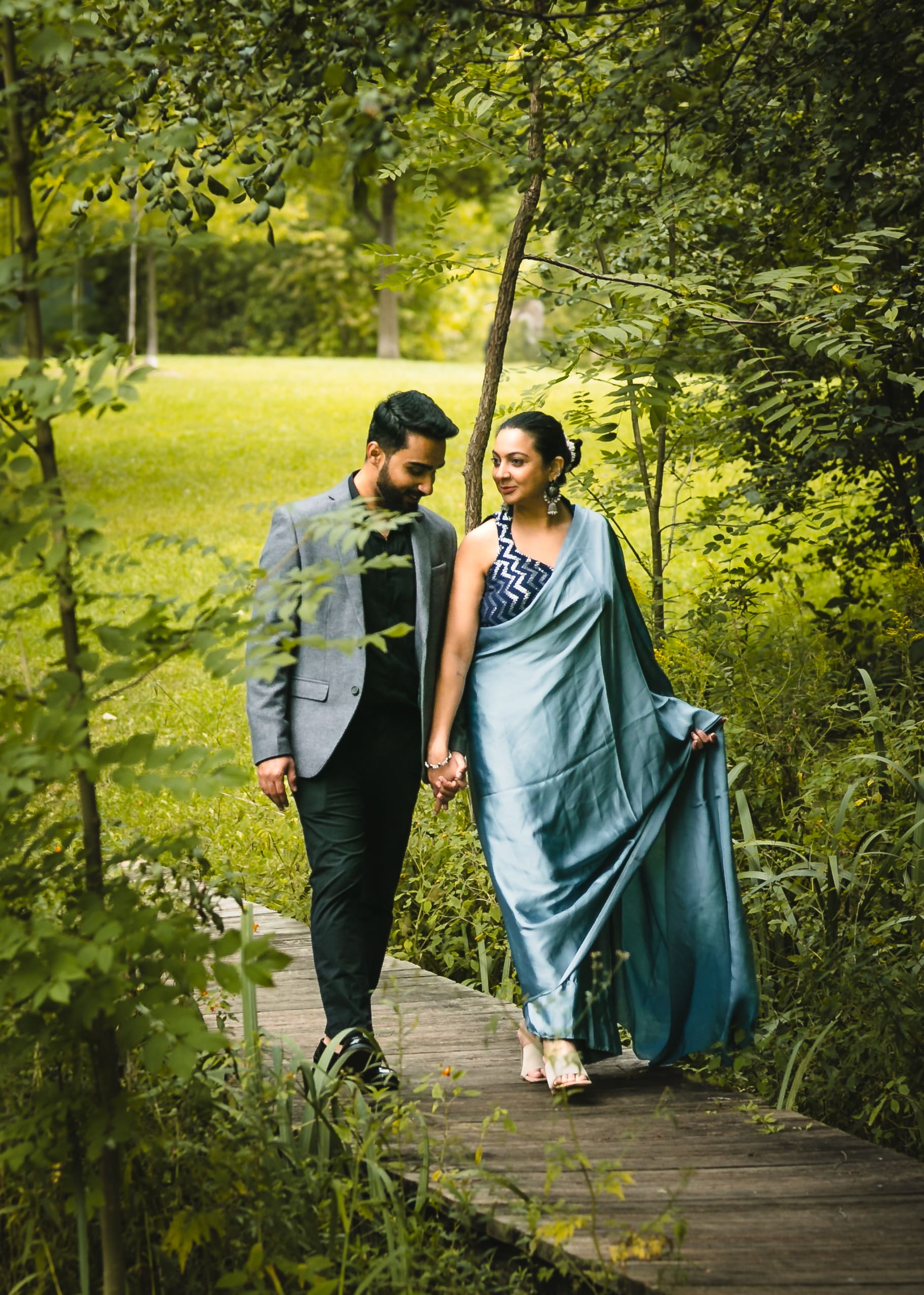 South Asian couple walking hand in hand on a wooden boardwalk in a lush green park, bride in a blue sari and groom in a grey blazer.