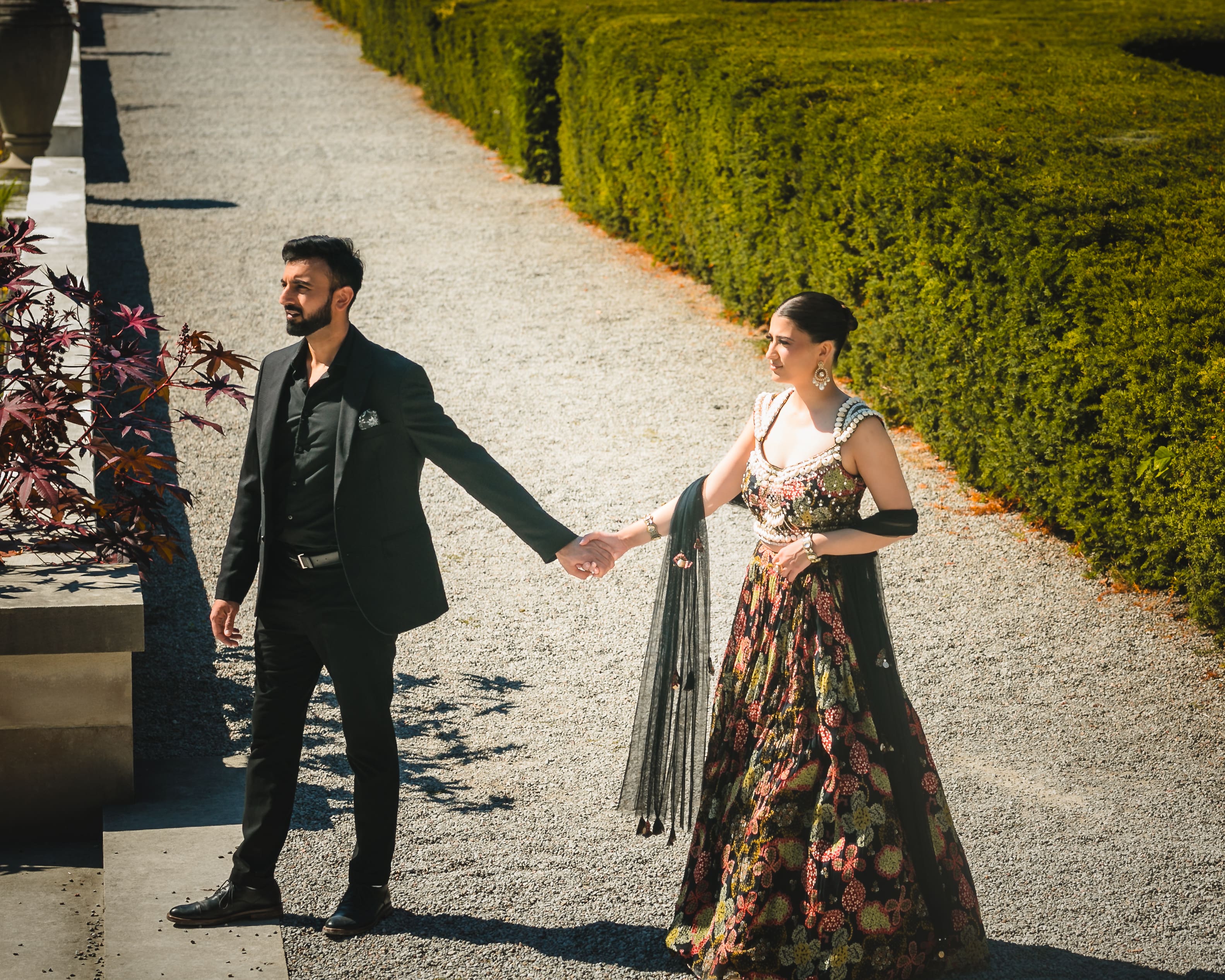 Wedding couple holding hands while walking along a garden path at Parkwood Estate in Oshawa, Ontario, bride in embroidered lehenga and groom in black suit, cinematic estate wedding portrait near Toronto and GTA.