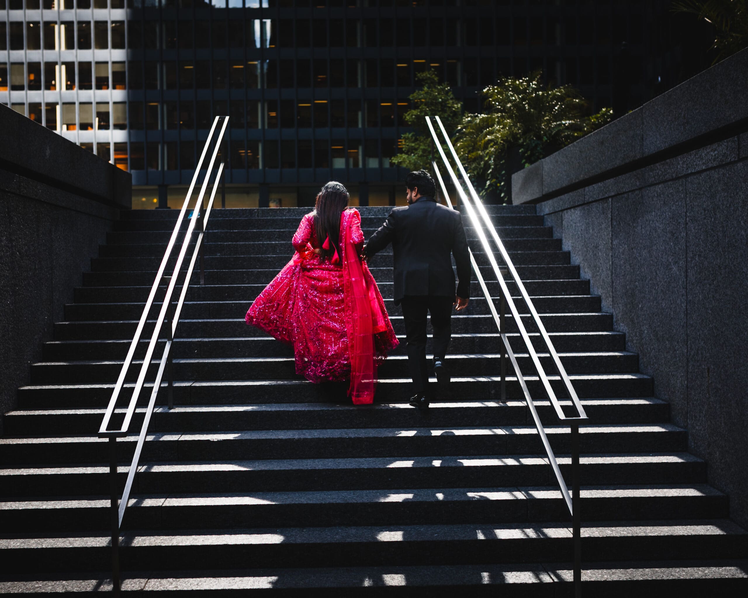 Downtown Toronto engagement photo of a couple walking up modern stone stairs, with the bride-to-be in a vibrant red outfit and the groom in a black suit, captured in a cinematic storytelling style.