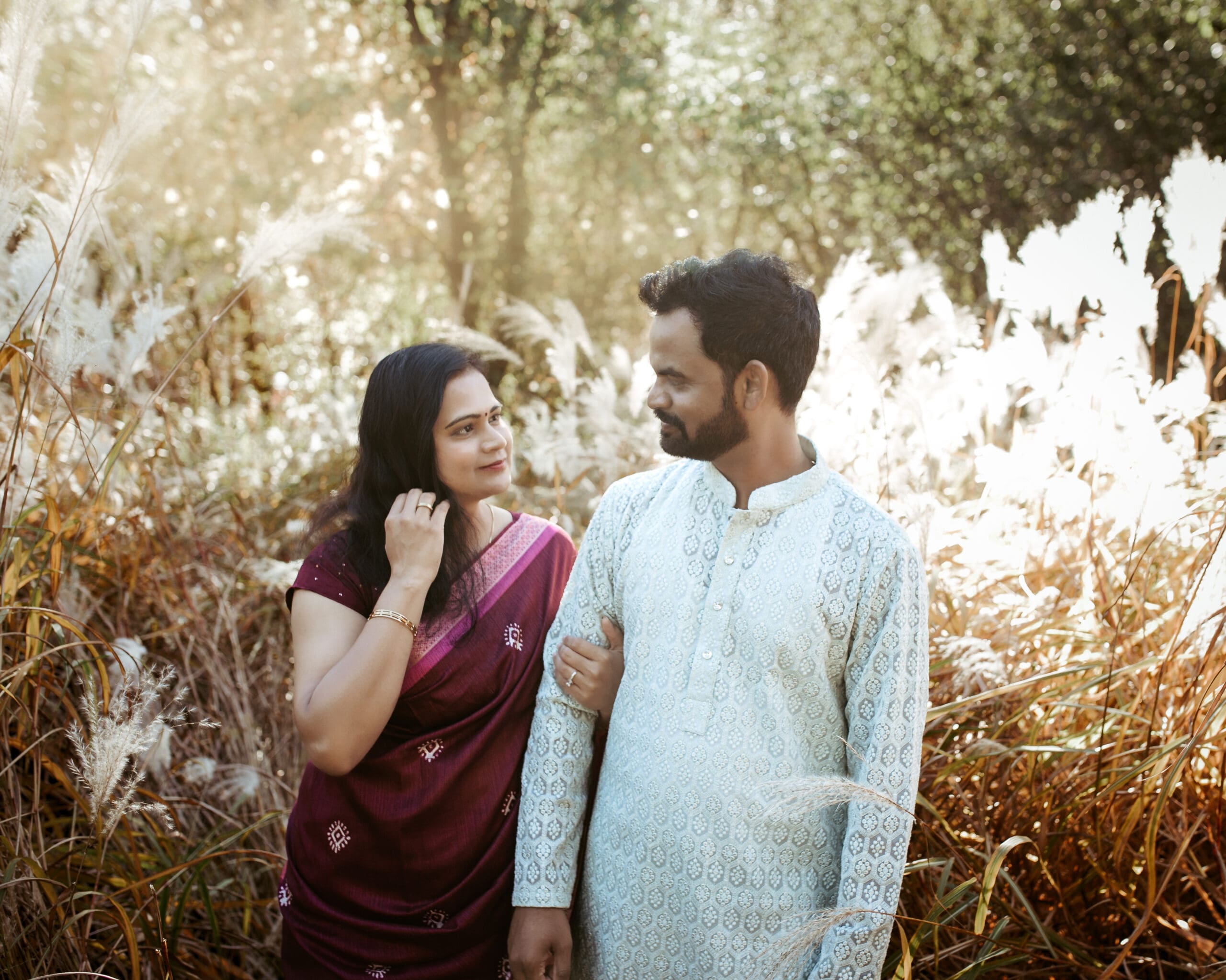 Toronto engagement photo of a South Asian couple in traditional outfits standing together in tall grass during golden hour, captured in a warm cinematic natural light style.
