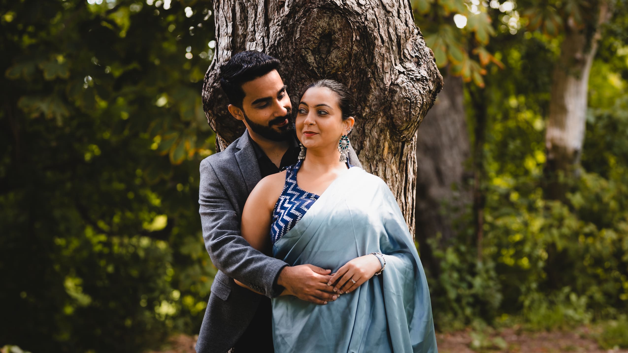 Toronto engagement photo of a South Asian couple embracing beside a large tree in a green park, with the bride-to-be in a blue sari and the groom in a grey suit, captured in a cinematic natural light style.