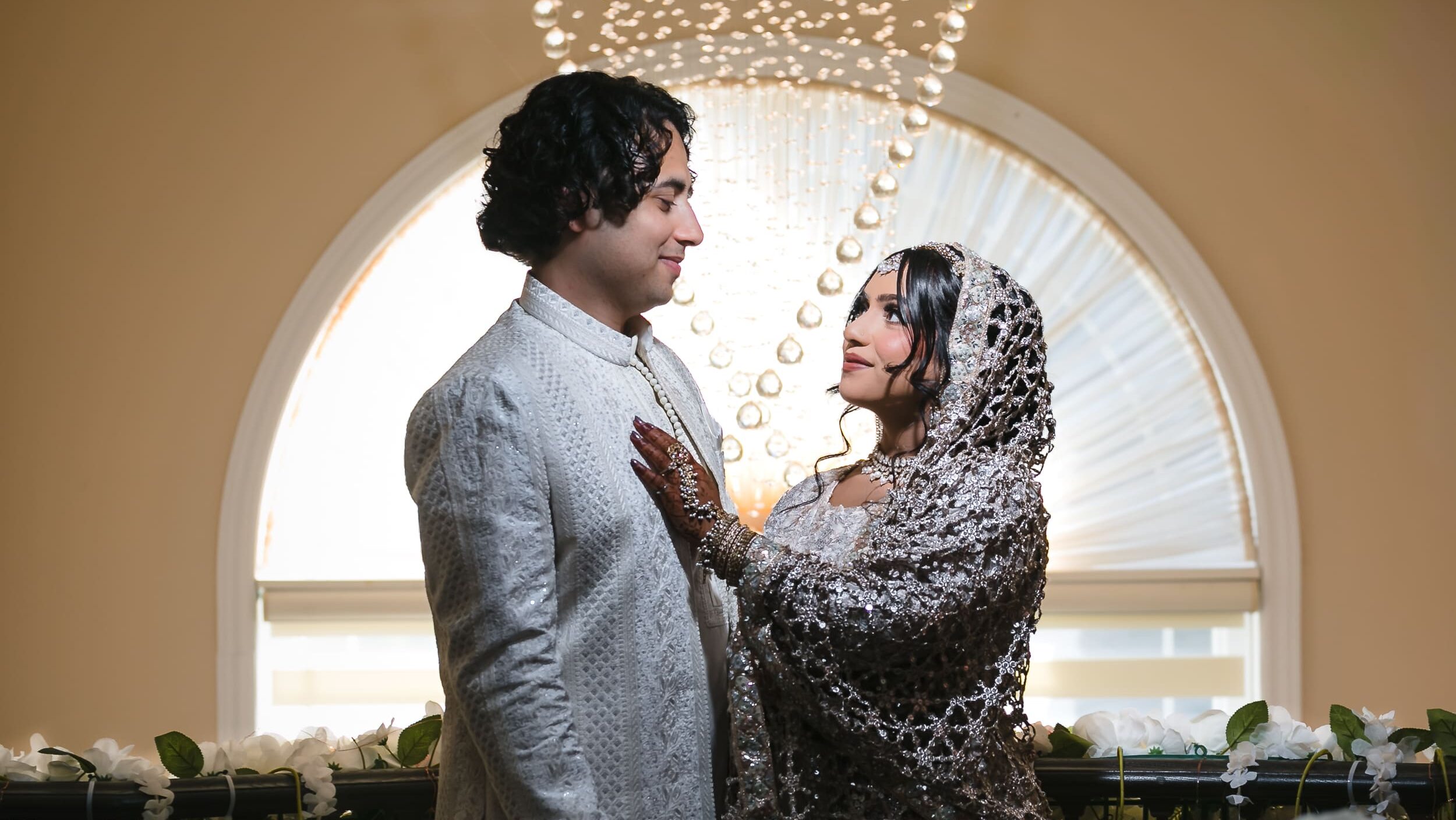 Bangladeshi couple sharing an intimate moment during their Nikah photoshoot in Toronto, standing beneath a chandelier beside an arched window and floral decorations