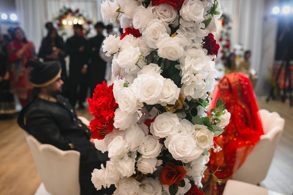 Close-up of white and red rose floral arrangement in foreground, with a groom in a black sherwani and turban seated to the left and a bride in a red bridal veil blurred in the background, during a Pakistani Nikah ceremony at Autaq Banquet Hall in Mississauga