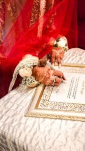 Bride's henna-decorated hand signing the Nikahnama marriage certificate, with a red bridal veil and cream floral hair jewelry in the foreground, against a deep red velvet backdrop