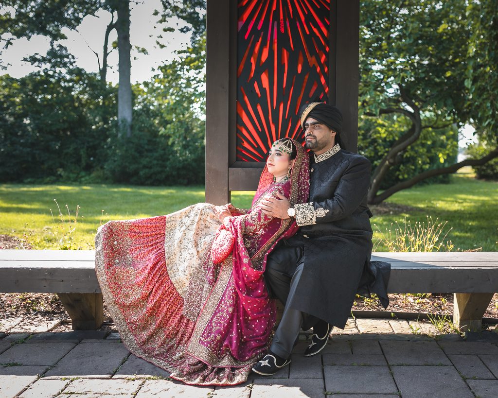 Pakistani couple in traditional wedding attire during an intimate Nikah photoshoot at Nottinghill Gate Park in Oakville, seated together on a wooden bench surrounded by greenery