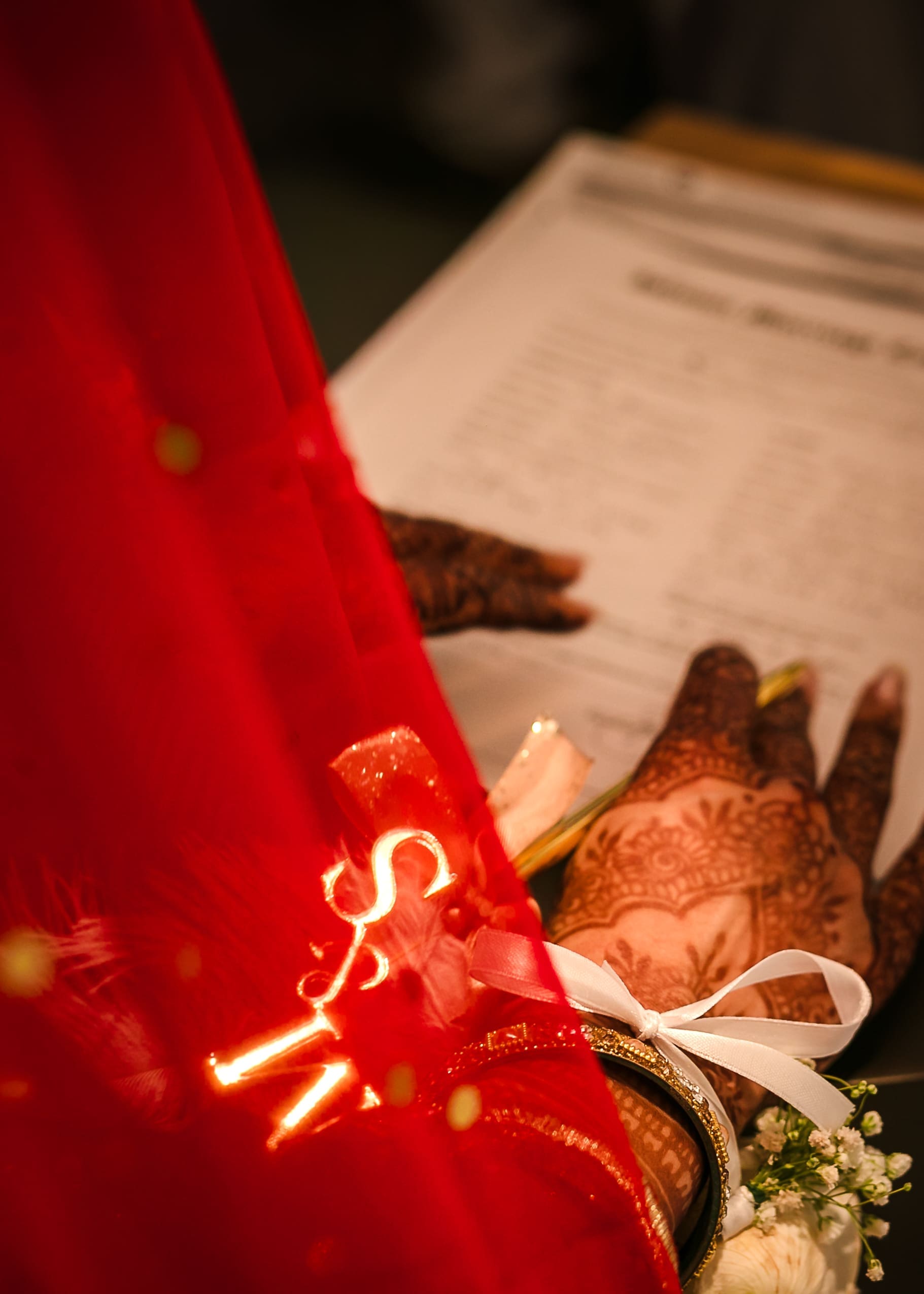 Close-up of the bride's henna-decorated hands resting on the Nikahnama marriage contract, with a red bridal veil, gold bangle, white ribbon bow, and baby's breath flowers in the foreground, during a Pakistani Nikah ceremony