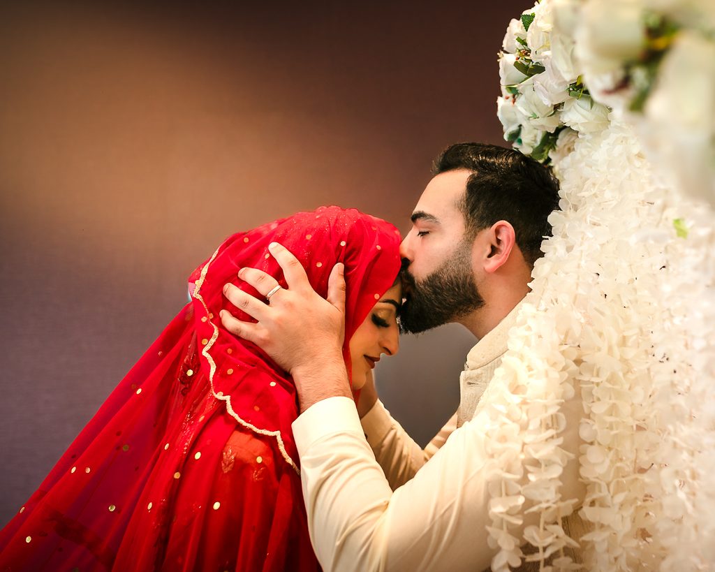 Groom in a white sherwani with a white floral garland kissing the bride's forehead as she wears a red bridal hijab and veil, during a Pakistani Nikah ceremony in Vaughan