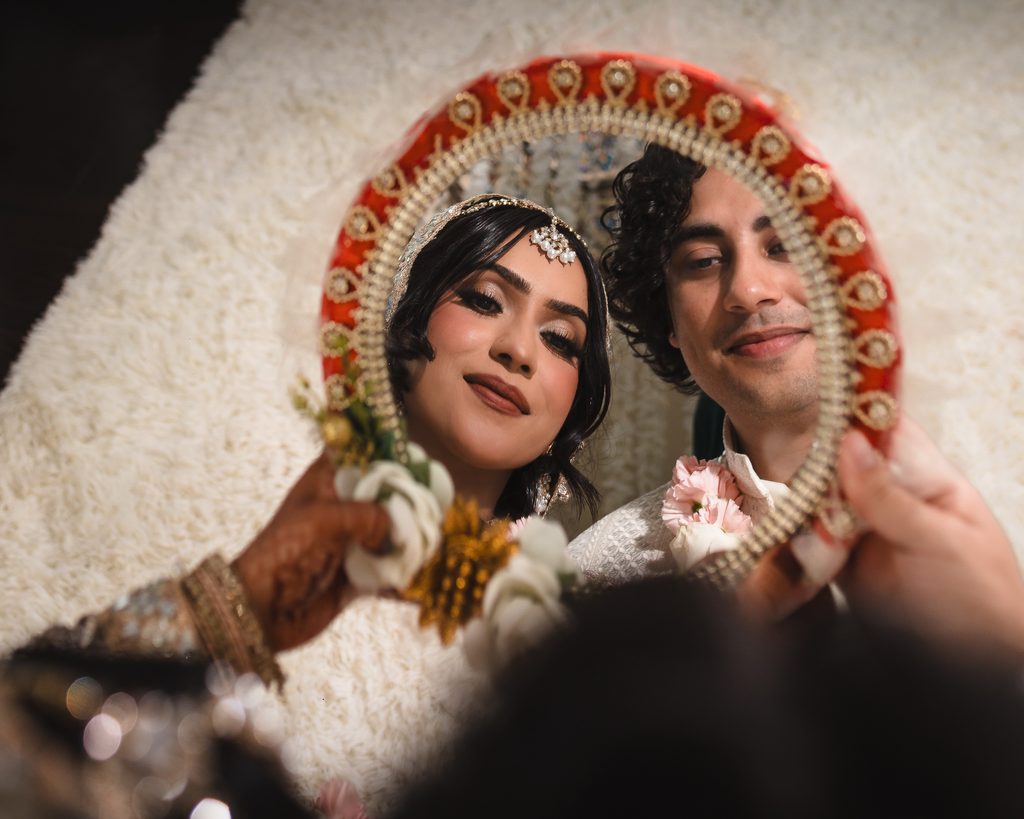 Bangladeshi bride and groom smiling into a decorative red and gold mirror held up to the camera, showing their reflection surrounded by a jeweled floral border, during their Nikah ceremony