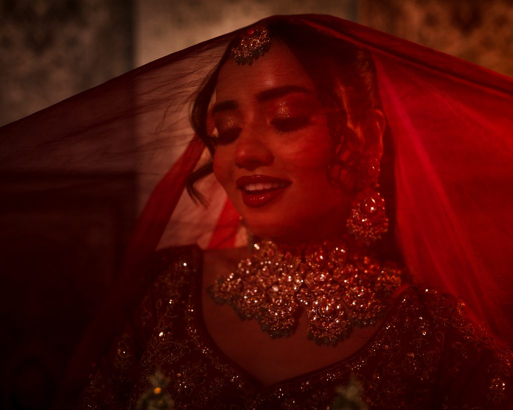 Bangladeshi bride in Mississauga smiling with her eyes closed under a sheer red veil, wearing a maang tikka, large drop earrings and a layered gold choker with green bead drops over her red embroidered outfit.