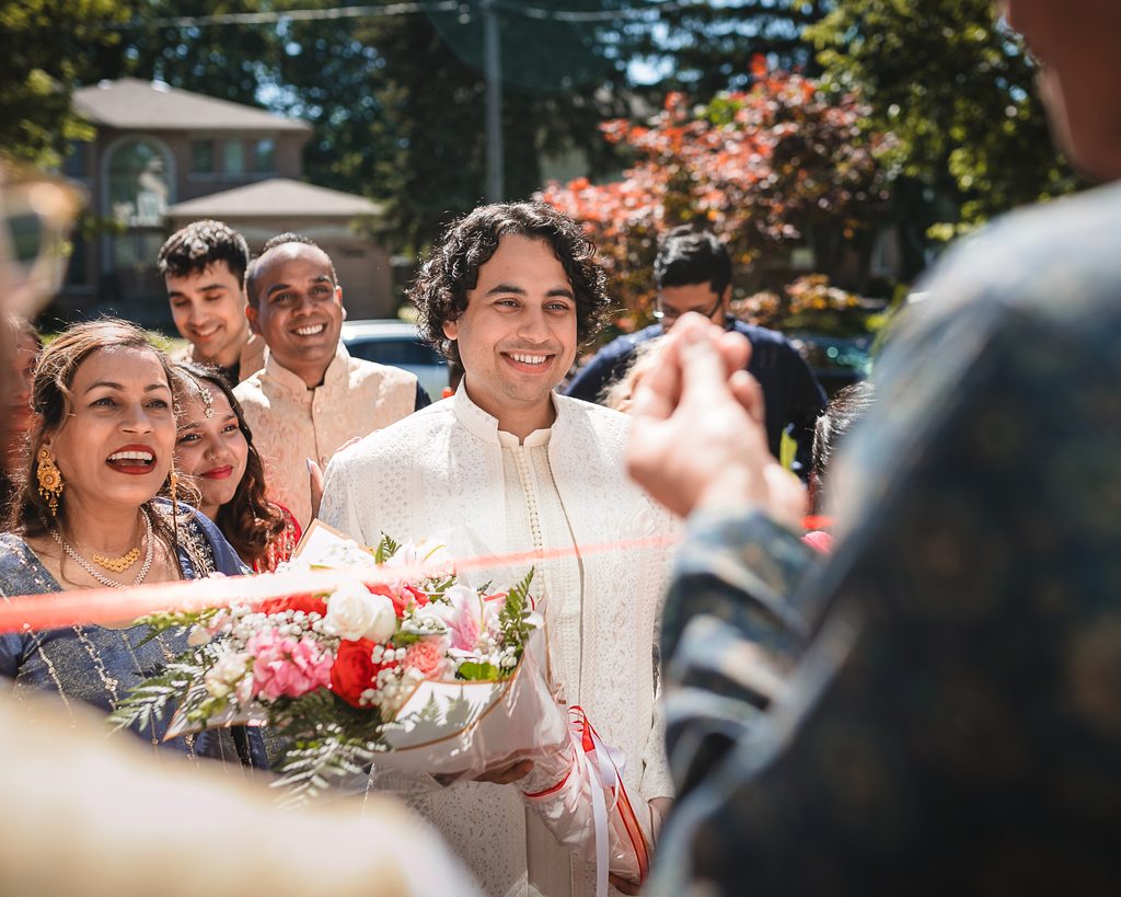 Smiling groom in a white embroidered sherwani holding a bouquet behind a red ribbon during gate dhora, with his family laughing beside him outside a home in Scarborough.