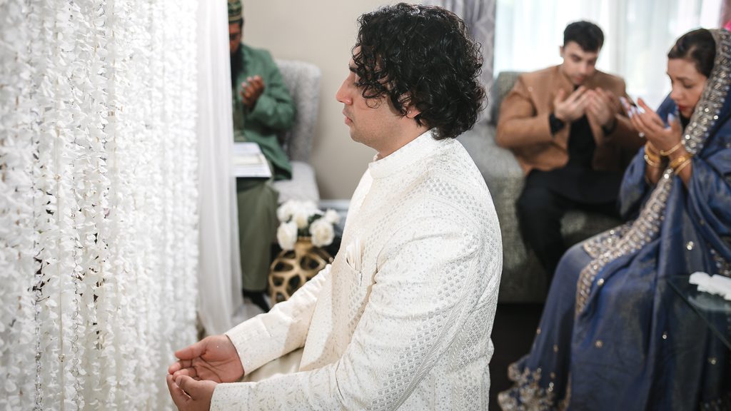 Groom in a white embroidered sherwani raising his hands in dua beside a white flower curtain during a Bangladeshi Nikah at home in Scarborough, with the Mawlana and family praying behind him.