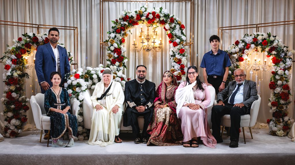 Bride and groom seated on stage with elders and family beside them, under a red and white floral arch with a chandelier, after a Pakistani Nikah at Autaq Banquet Hall in Mississauga.