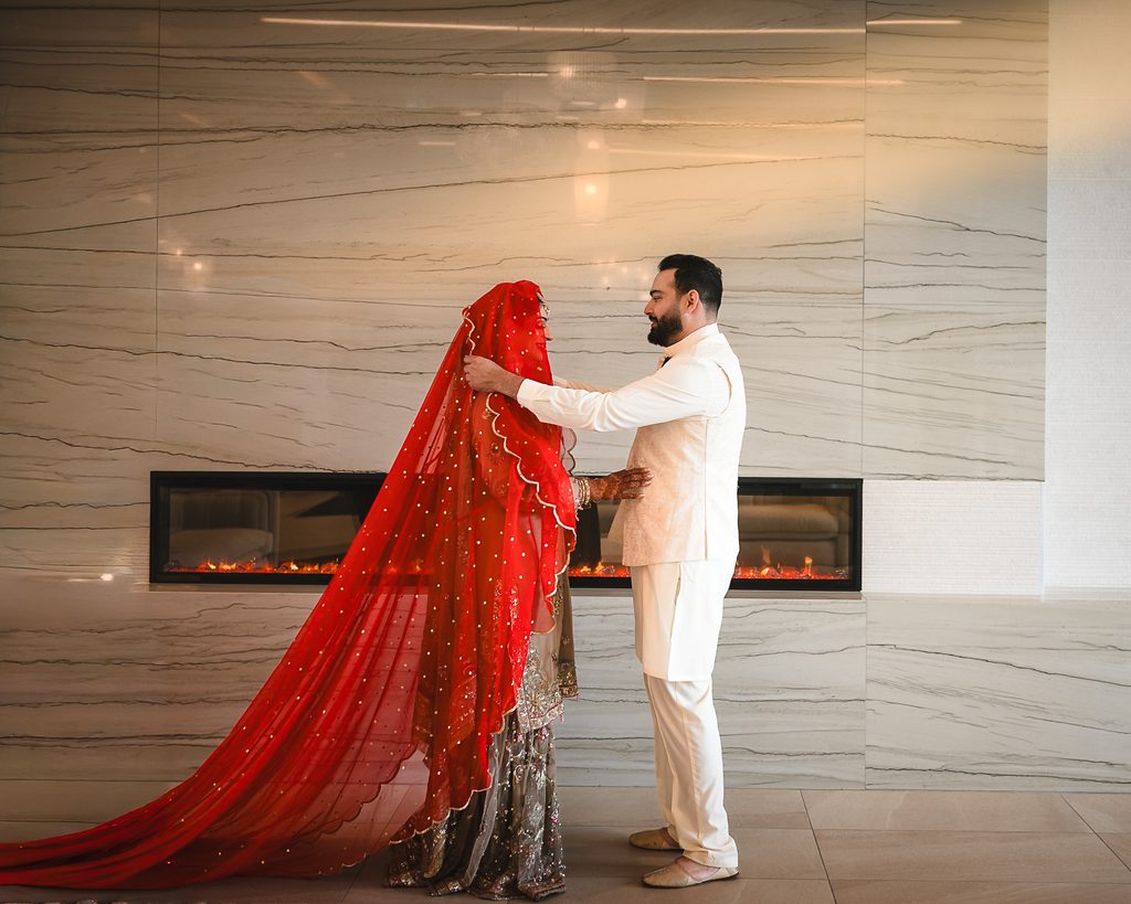 Groom in a white kurta and cream waistcoat lifting the bride’s long red sequined veil as she rests her hennaed hand on his side, in front of a marble wall and fireplace at a Pakistani Nikah in Vaughan.