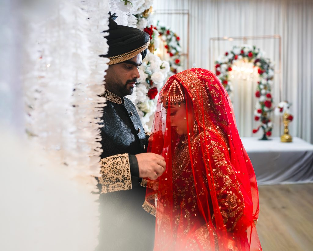 Groom in a black sherwani and turban with gold embroidery holding the edge of the bride’s red embroidered veil beside a white flower curtain during a Pakistani Nikah at Autaq Banquet Hall in Mississauga.